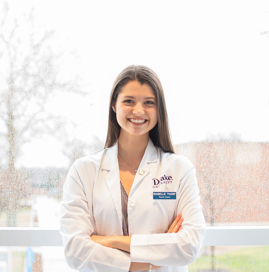 Student Isabelle Tharp stands in front of a window on campus while it rains behind her, with a white coat on.