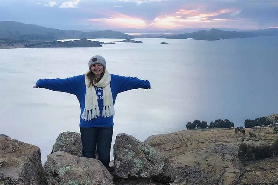 During a study abroad trip, a student in a Drake sweatshirt, beanie, and scarf spreads their arms out in front of a lake