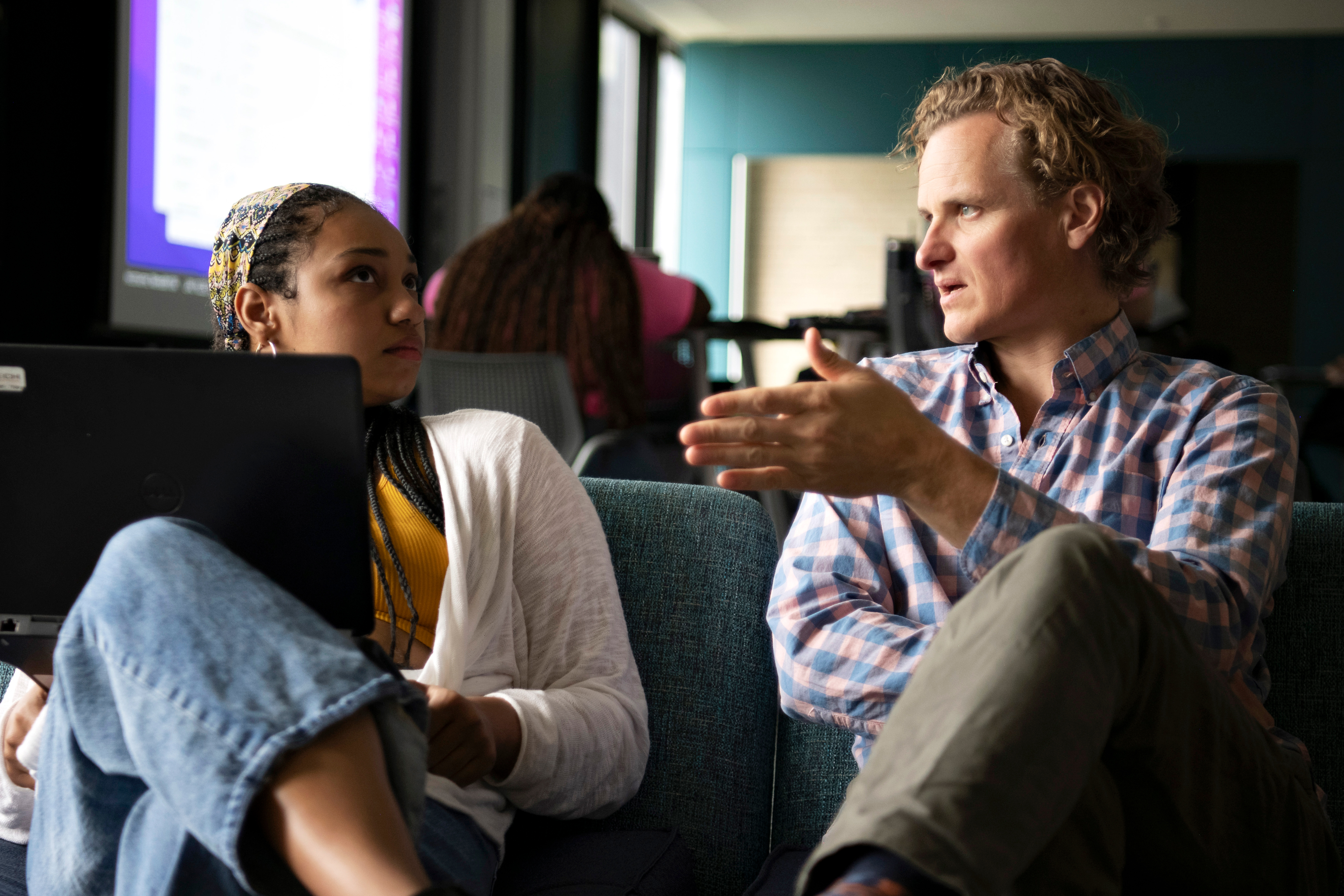 A faculty member meets with a student, supporting their laptop with one leg, in a campus common area