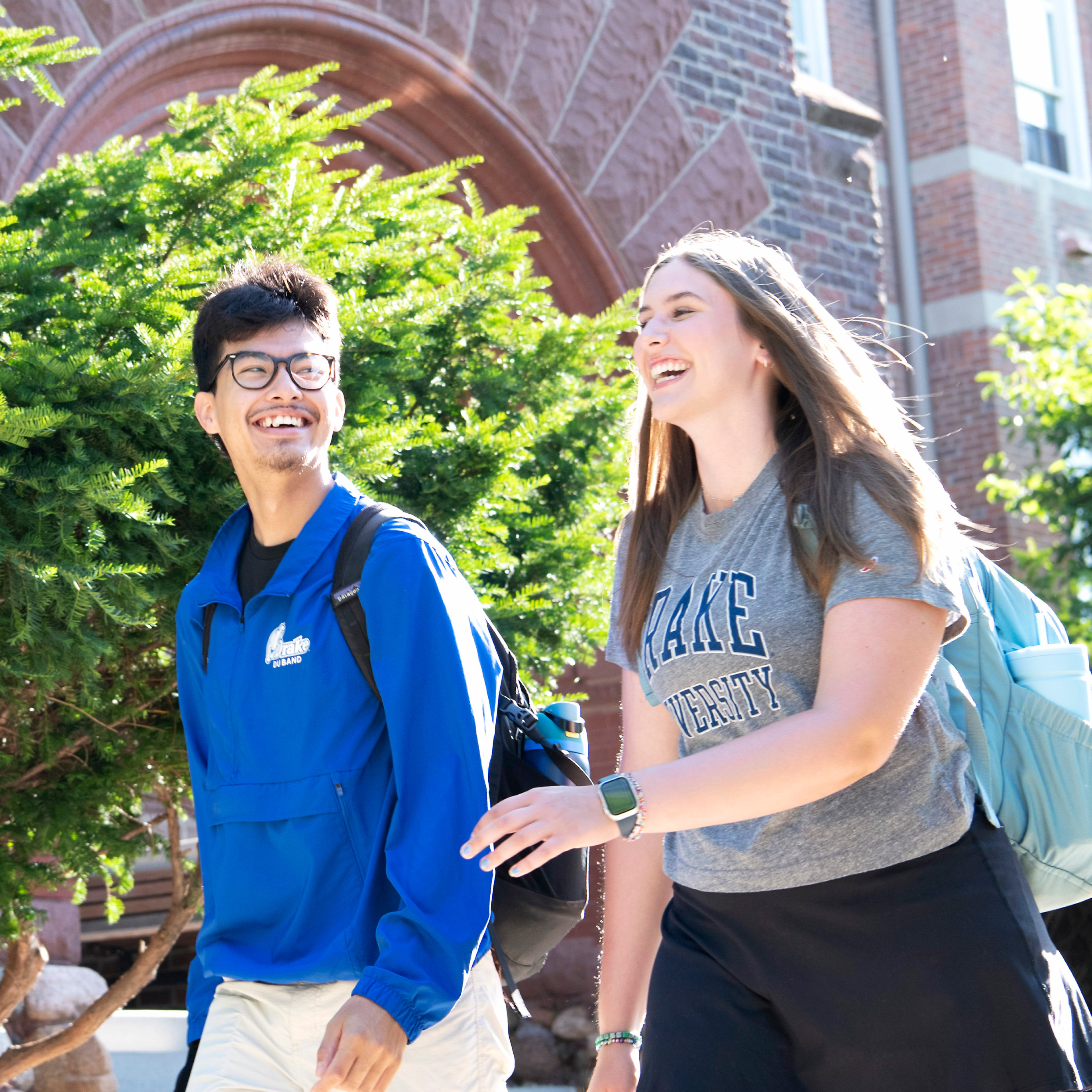 Two students, one in a school T-shirt, walk by a brick arch and a tree on the Drake campus