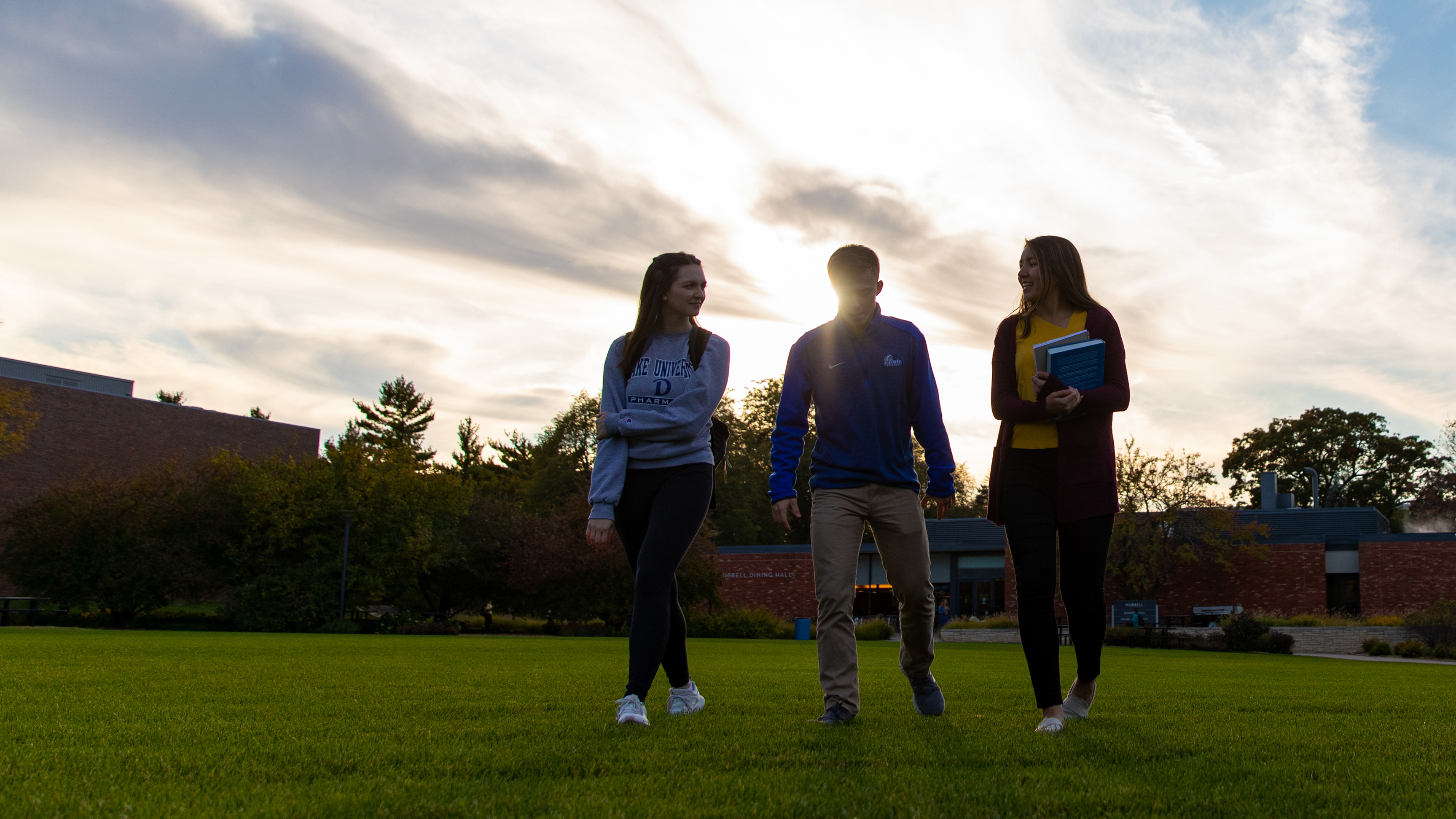 Three Drake students amble across a field, the sun setting behind buildings in the background