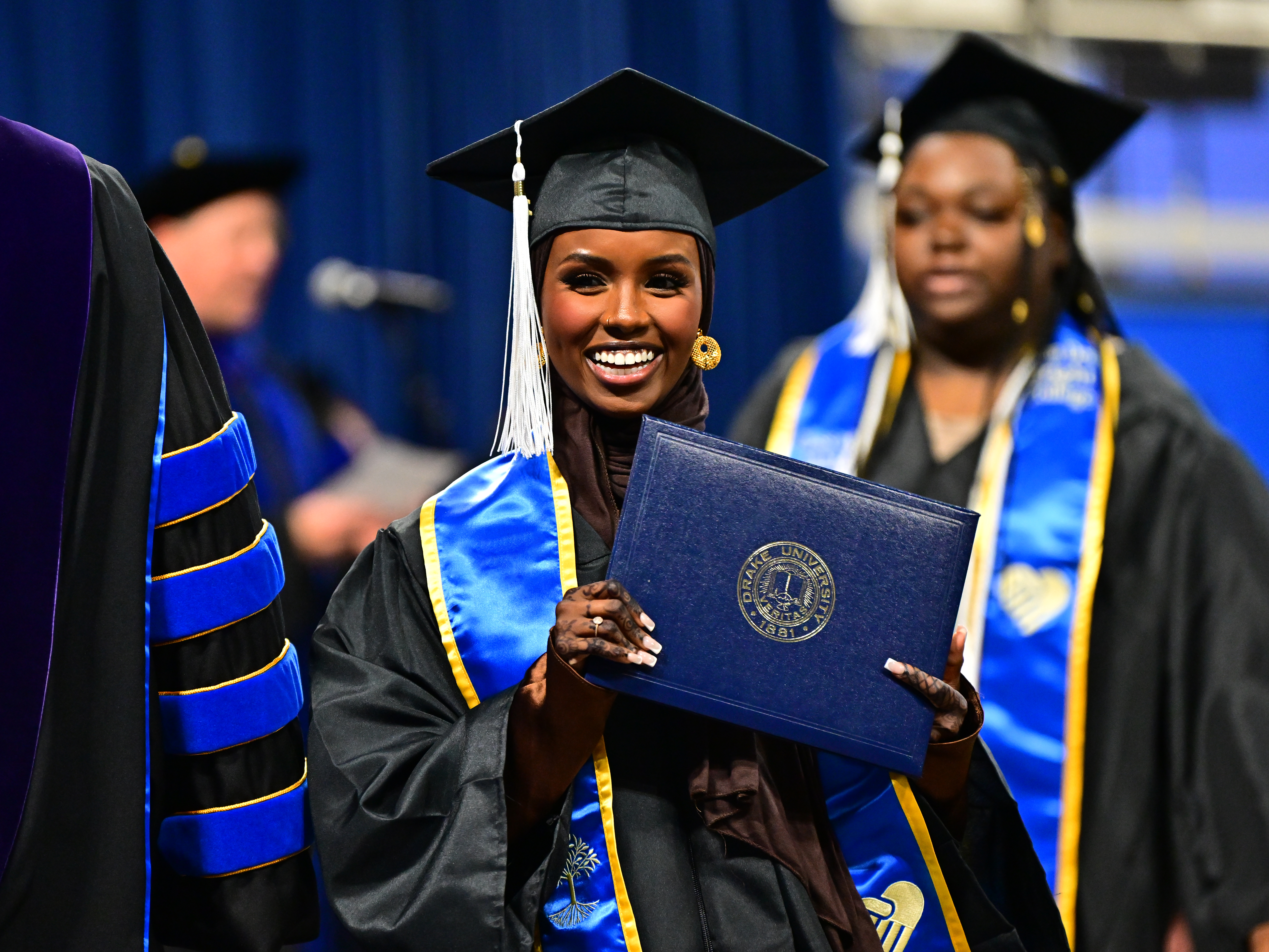 Walking on state during commencement, a student in a cap and gown smiles while holding their diploma