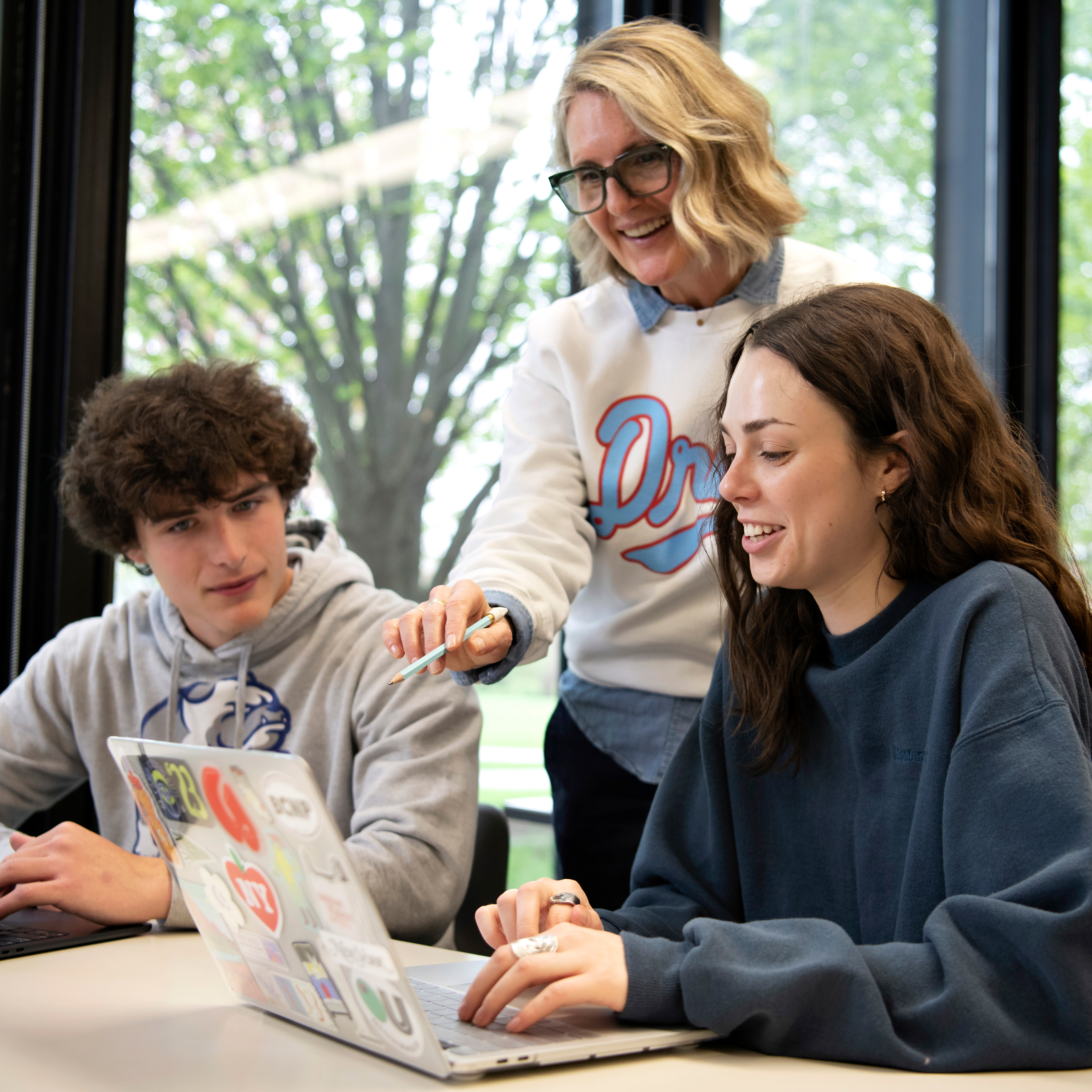 A Drake faculty member in a school sweatshirt points toward a student's laptop screen as another student leans to learn more