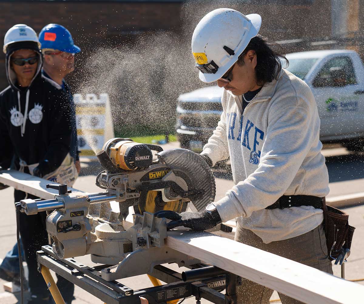Student in a hard hat using a miter saw while volunteering with Habitat for Humanity on Drake University's campus.