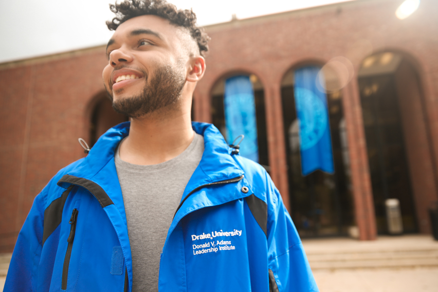 A student in a a blue jacket embroidered with Drake University, Donald V. Adams Leadership Institute, smiles while standing outside of a building on campus