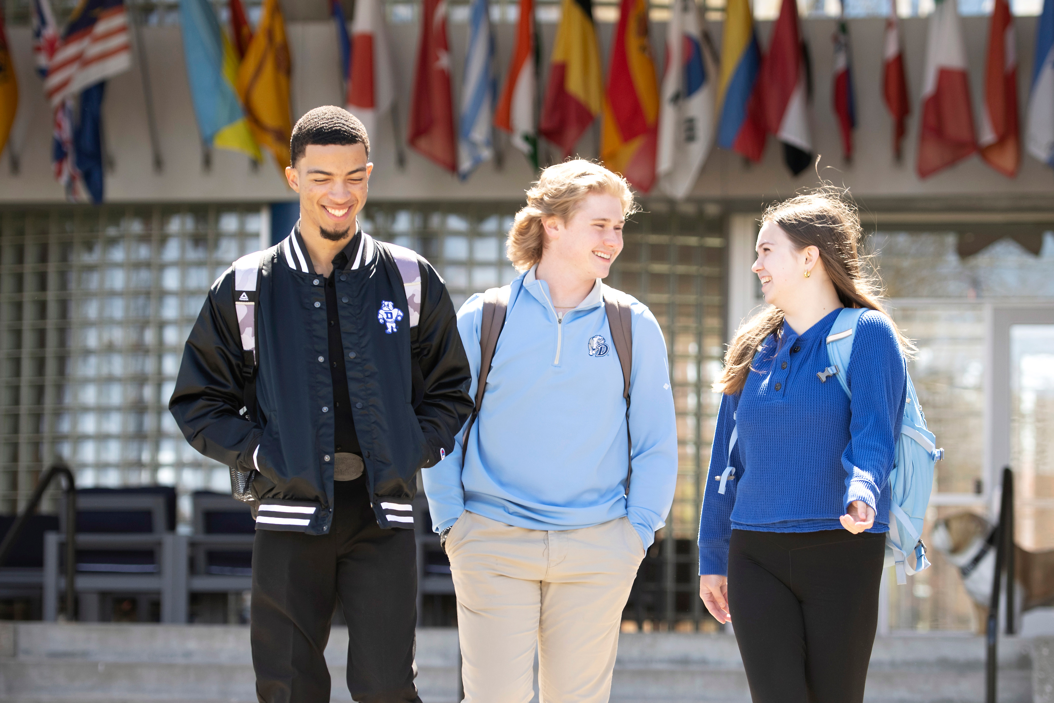 Three students with backpacks depart Drake's Zimpleman College of Business, a building featuring international flags