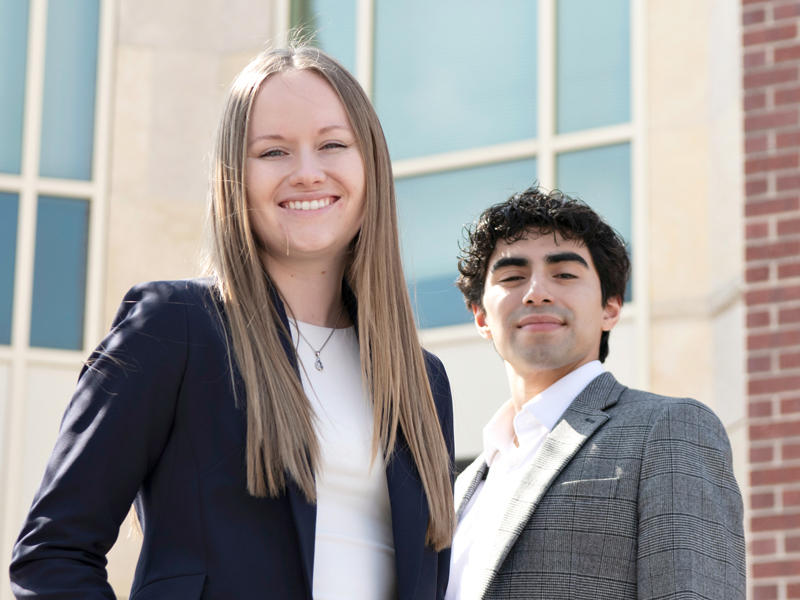 Two students in business attire smile while standing outside in front of a campus building