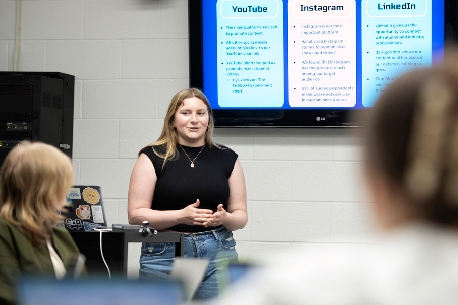 A student stands in front of a classroom, speaking, while a presentation is shown on the screen in the background