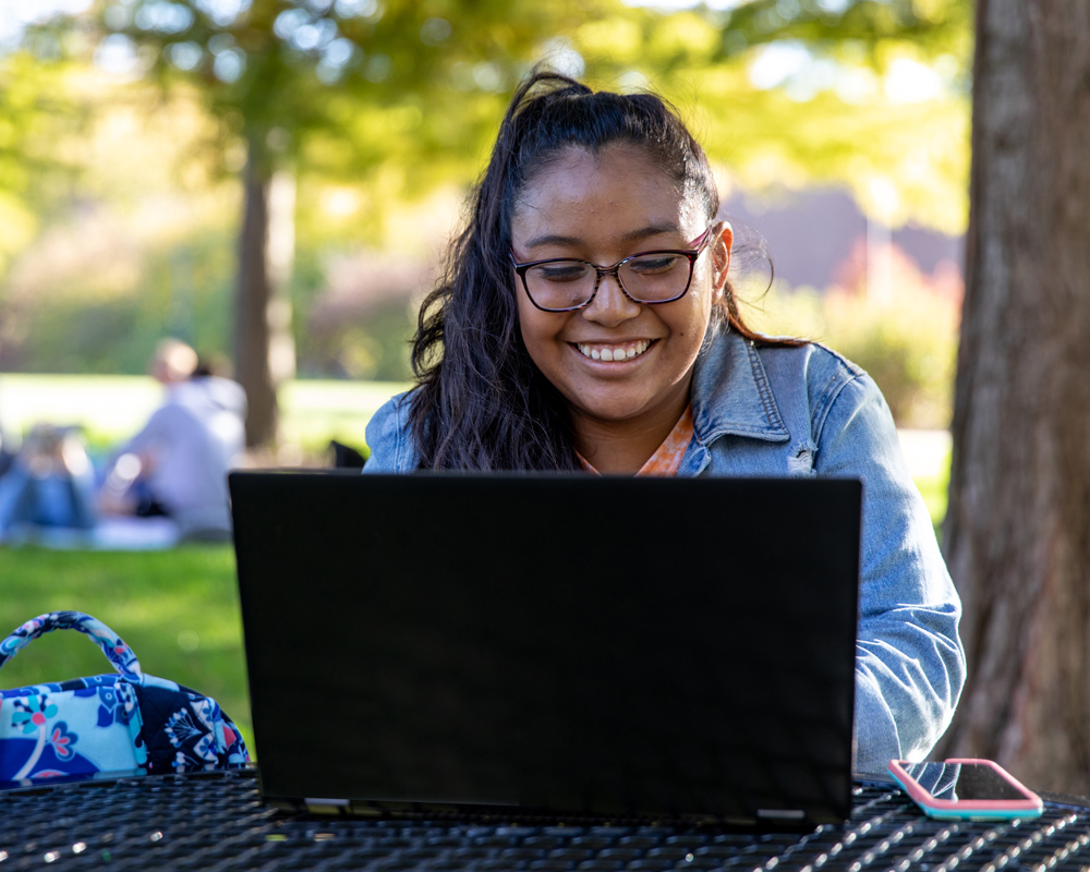 A student smiles while looking at a laptop and sitting outside on campus