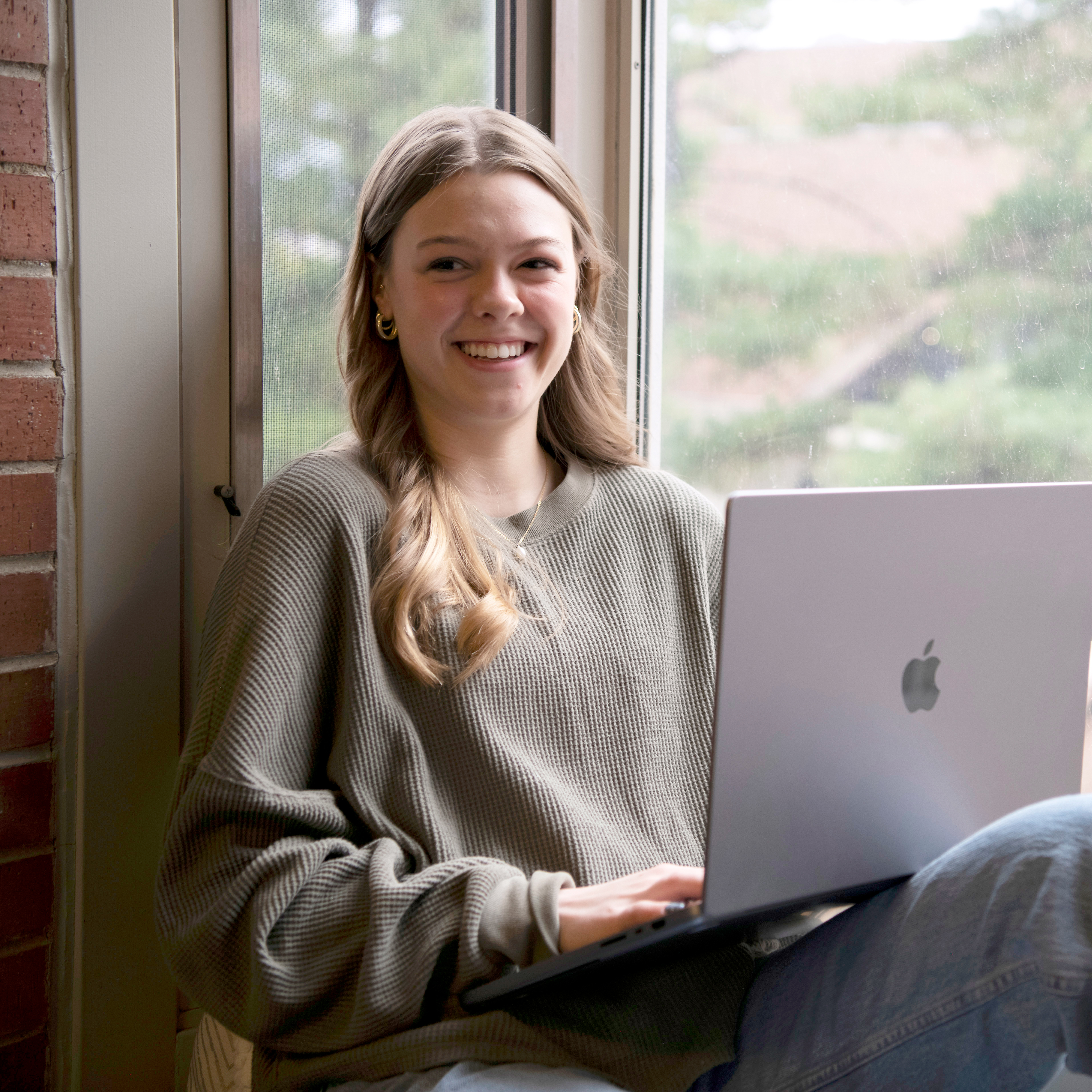 A Drake student smiles while sitting on a window ledge with their laptop