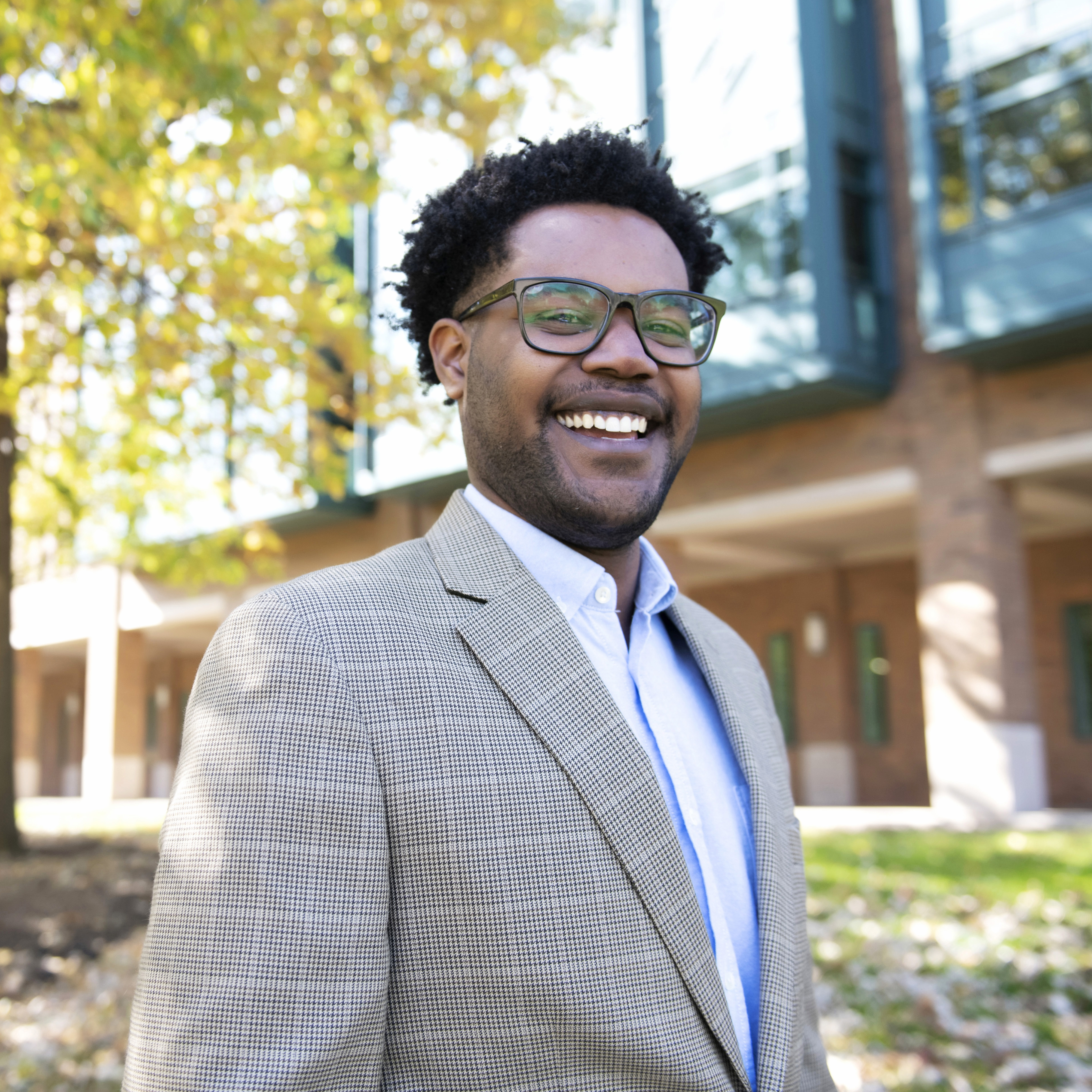 A Drake graduate student in glasses and a blazer smile while standing near a classroom building and trees