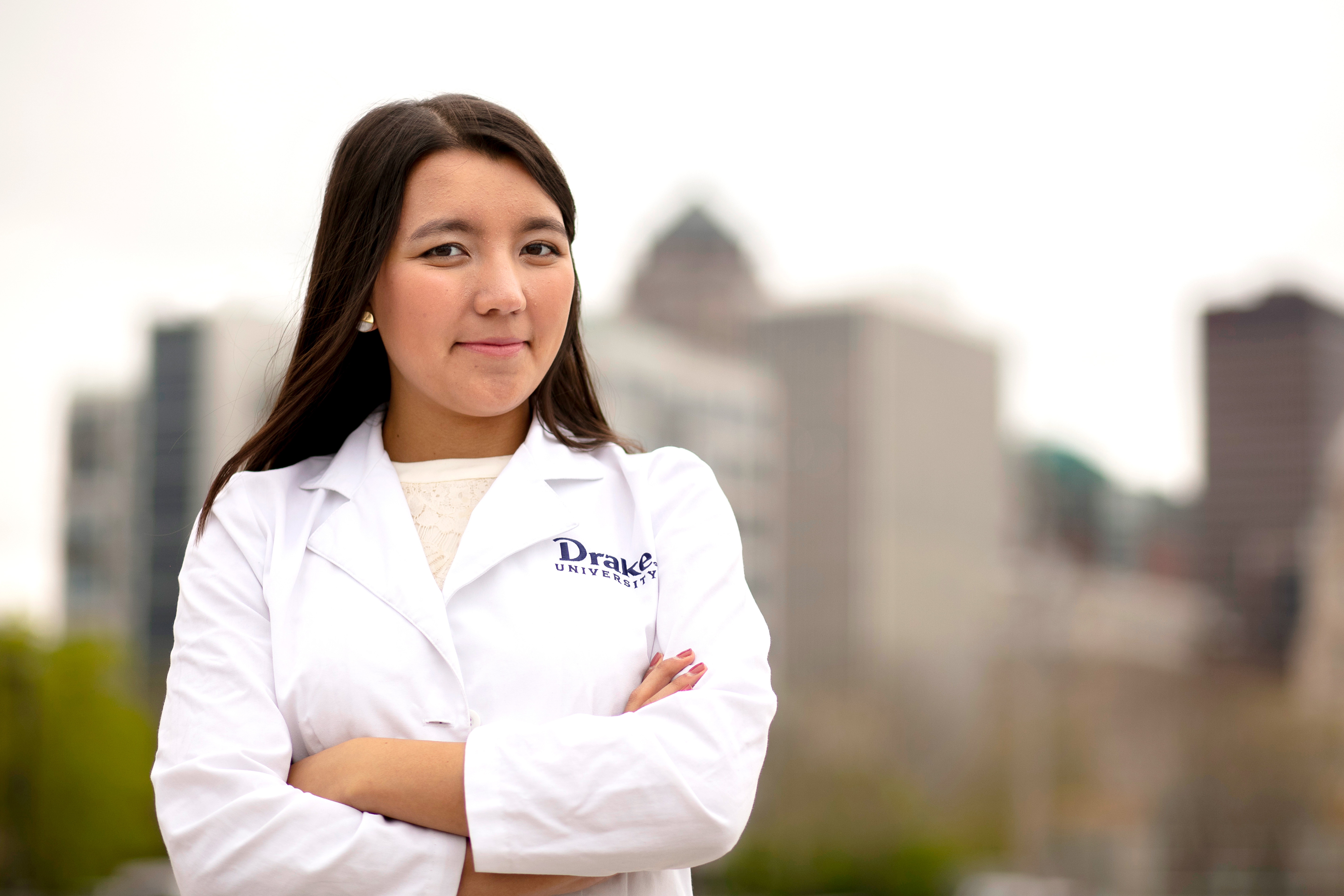 A international student in a lab coat with the Drake logo folds their arms. The Des Moines skyline is in the background