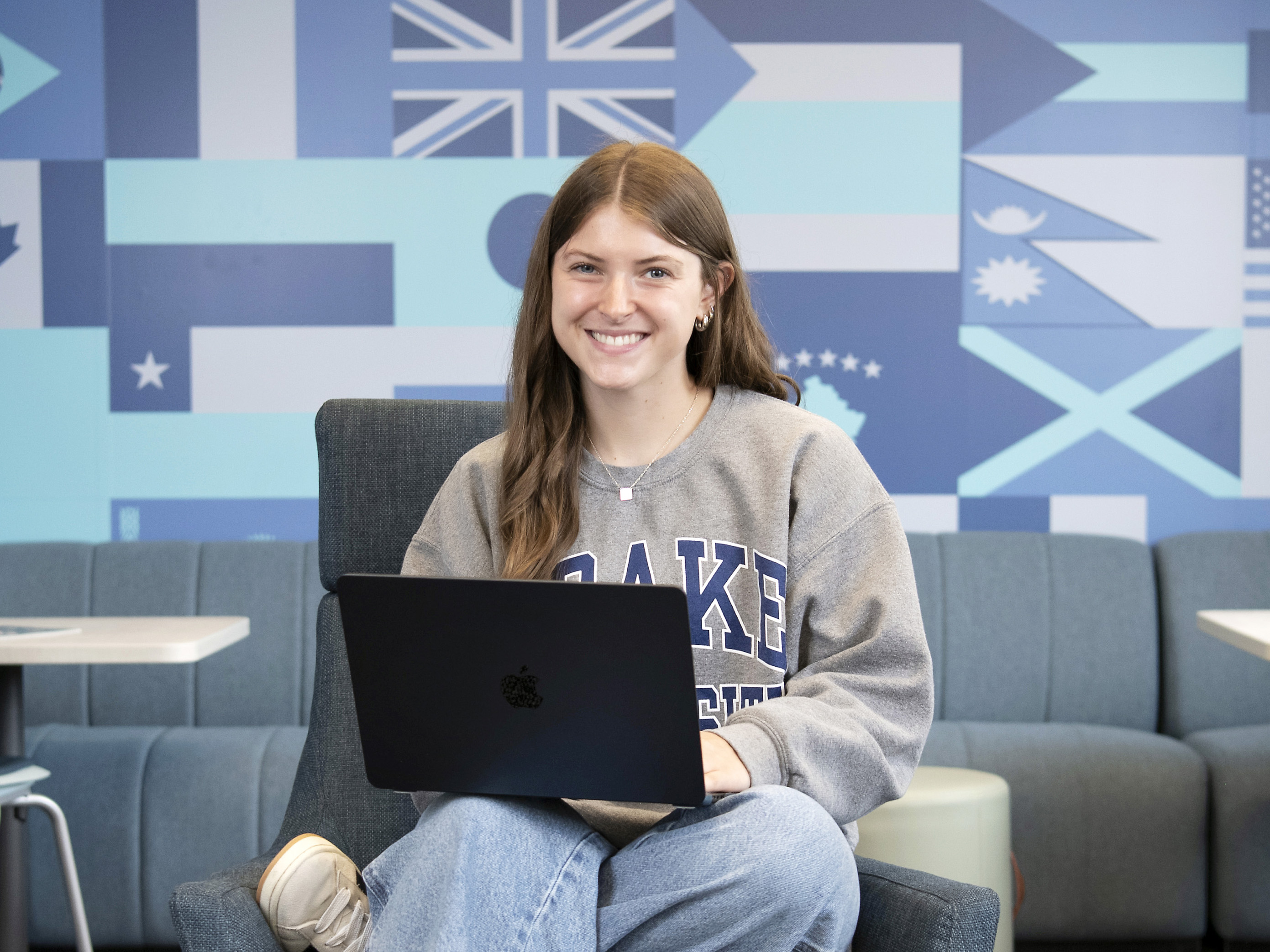 A Drake student supports their laptop on their lap while seated in front of a wall with blue- and gray-toned international flags