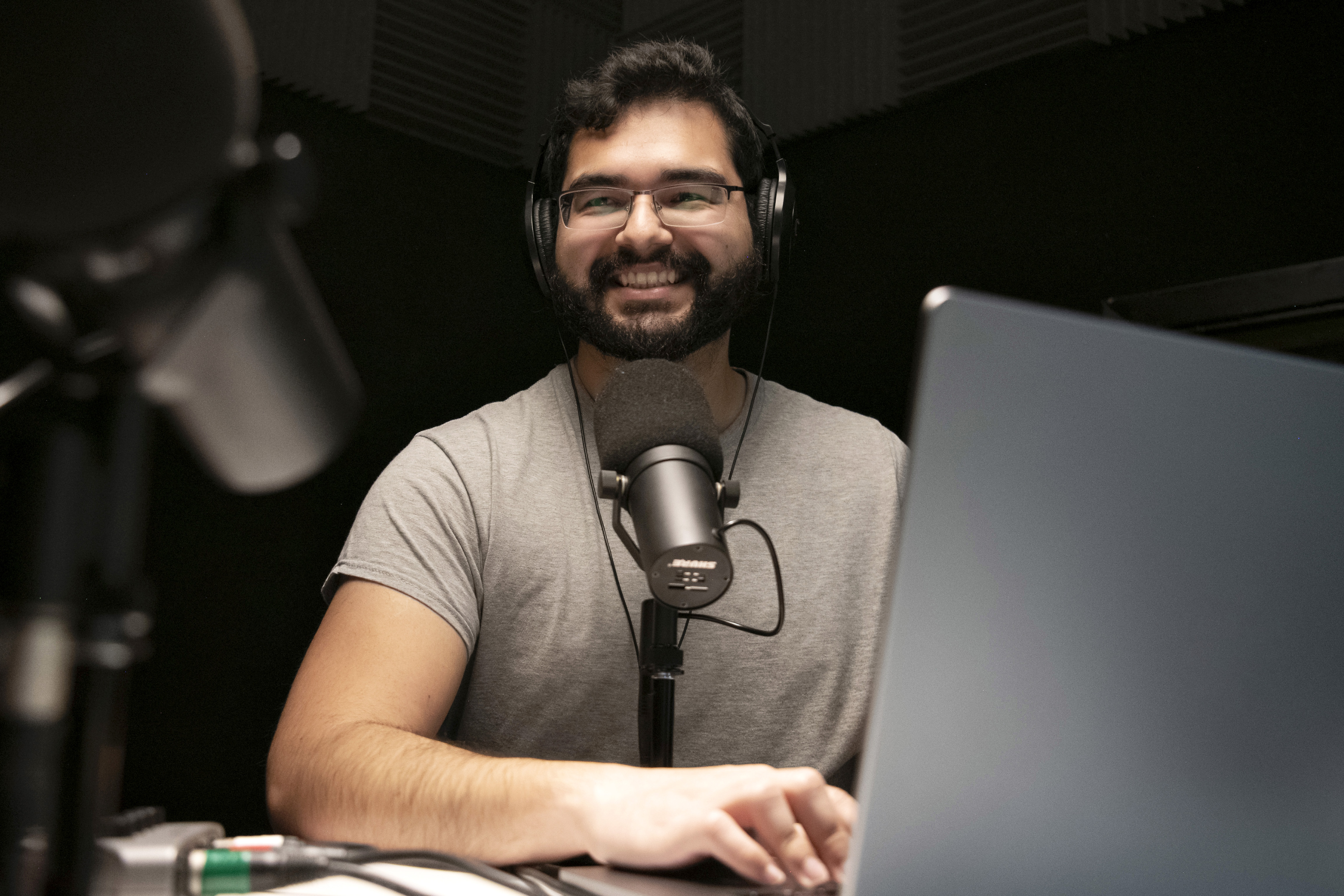 An international student with a microphones smiles from behind a laptop in one of Drake's media studios