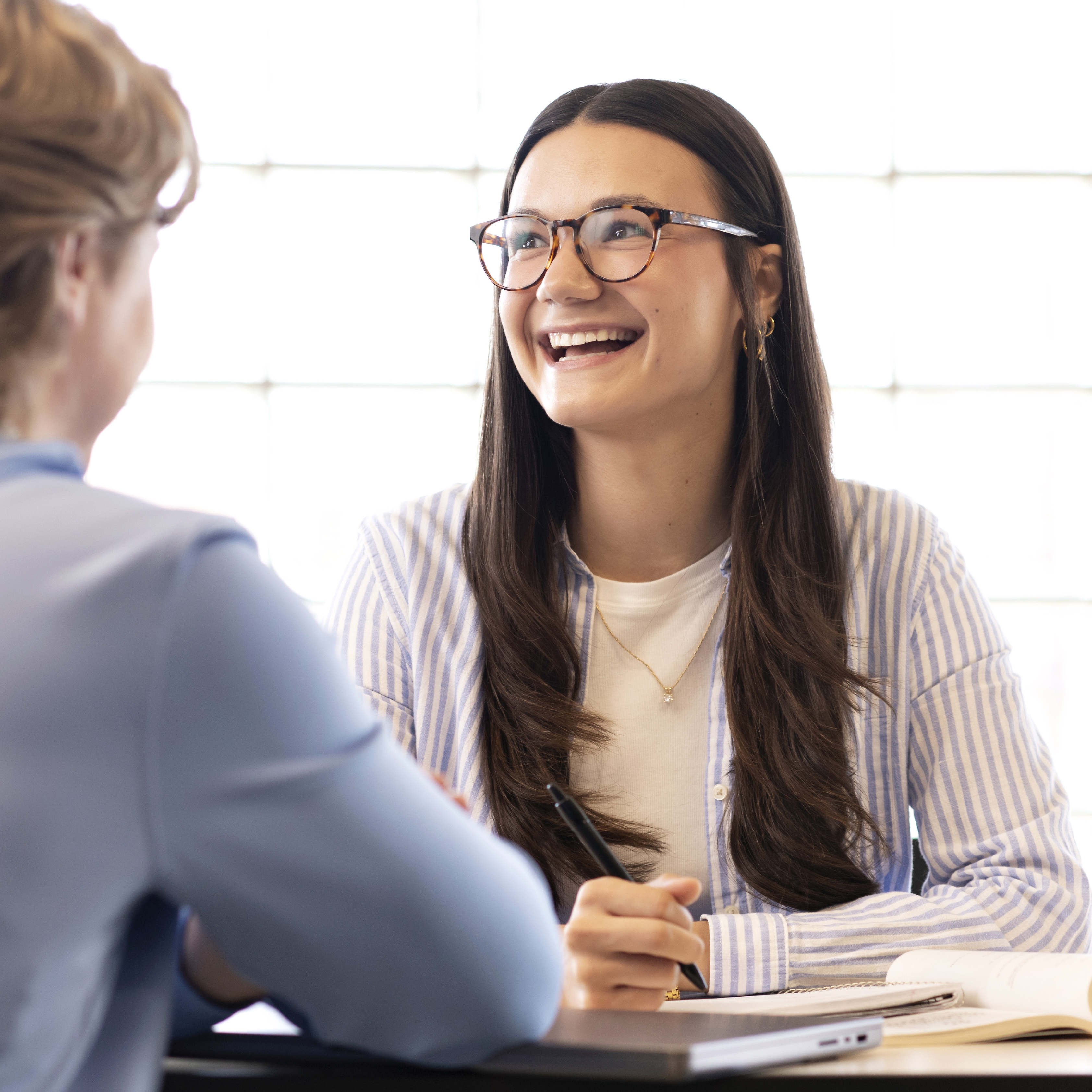 A Drake international student in glasses and a striped shirt smiles and slightly looks away during a conversation in a room lit with natural light