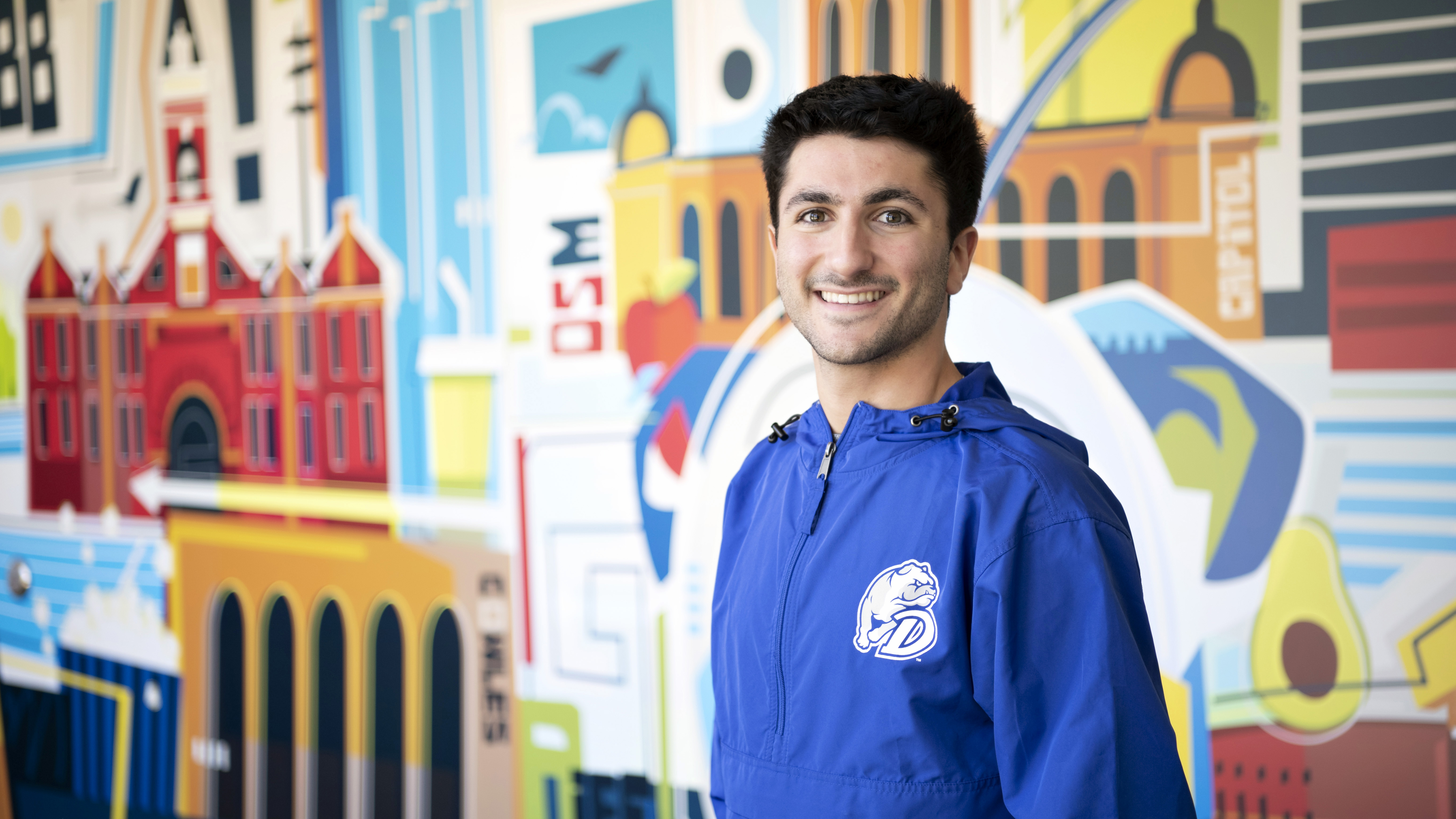A transfer student in a blue Drake anorak stands in front of a colorful mural on campus