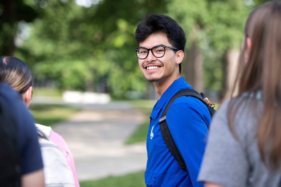 A student smiles at the camera while walking on campus with others who face away from the camera
