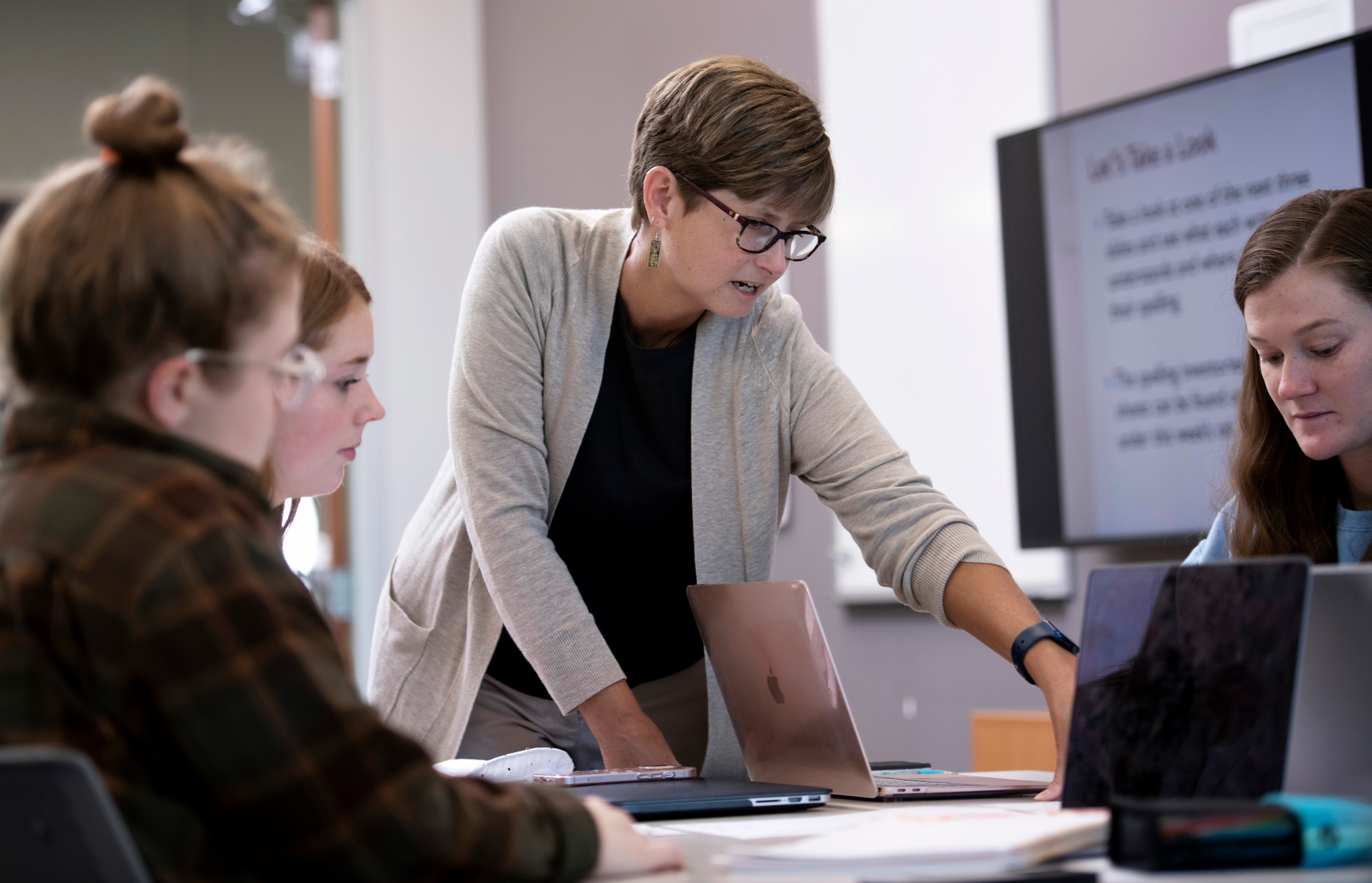 A professor stands over a table of students pointing at a laptop screen