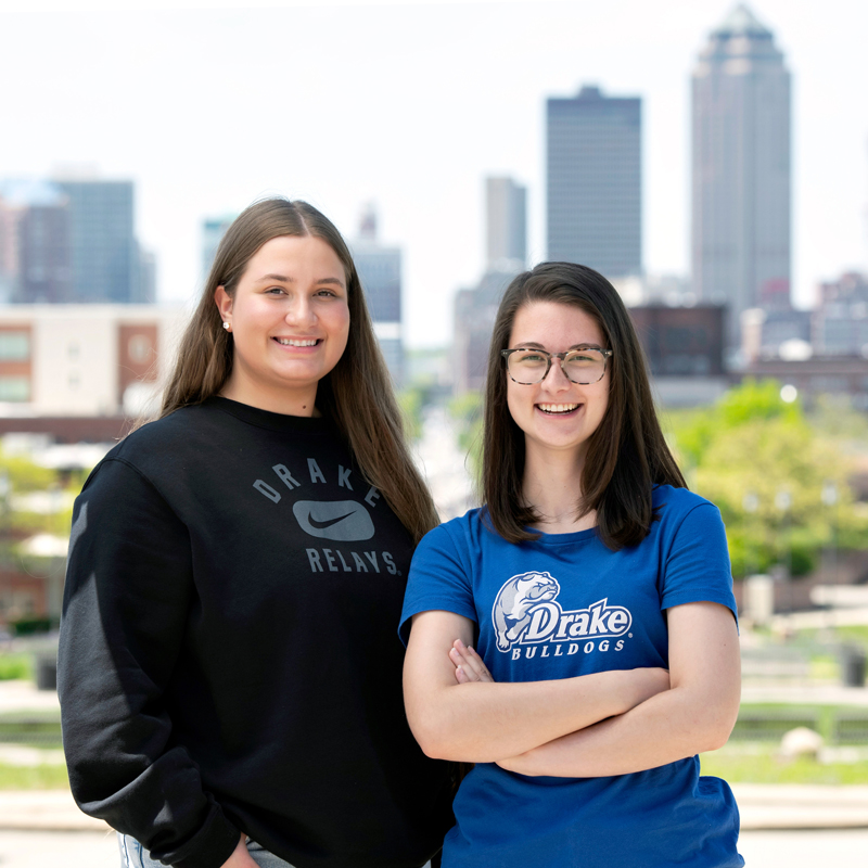 Two students stand, smiling, with a cityscape skyline in the background