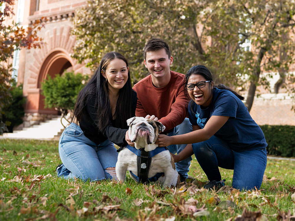 Three students sitting on the grass in front of Old Main during the Fall, with Griff II.