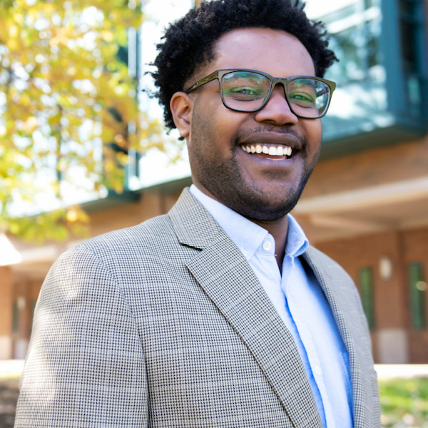 Smiling man wearing glasses and a gray checkered blazer, standing outdoors near a building with trees in the background.