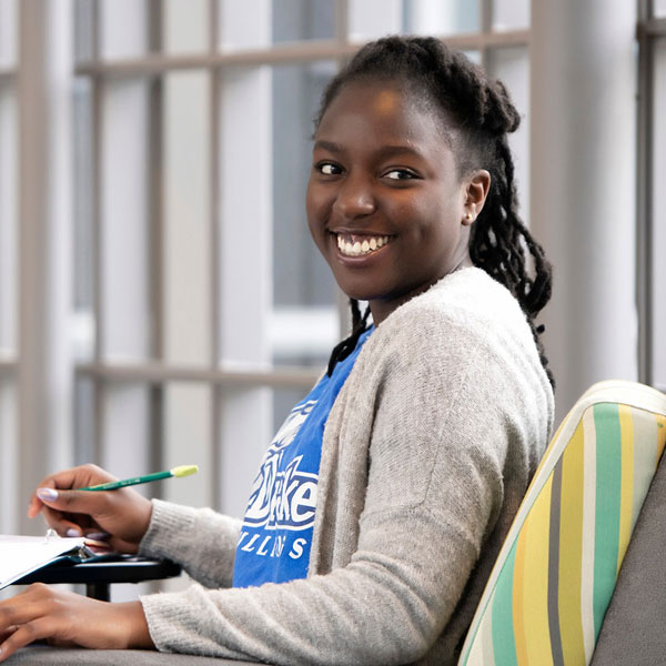 Smiling student seated indoors holding a pencil and notebook, looking toward the camera.