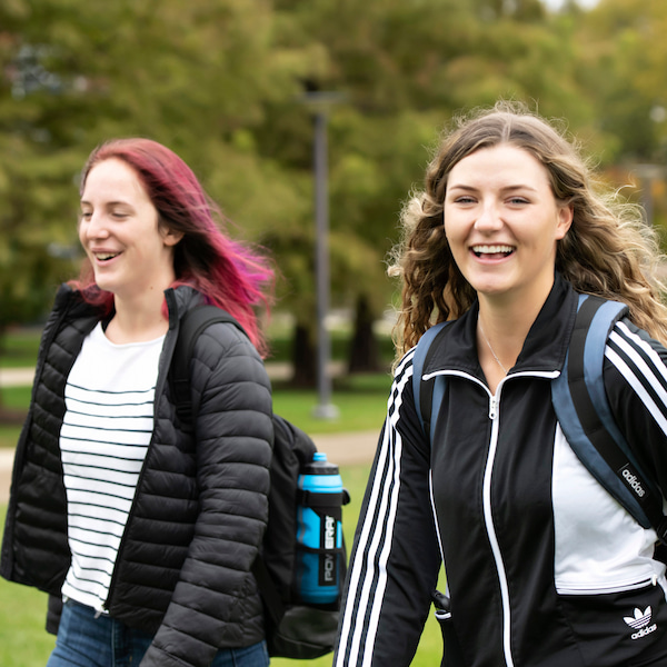 Two young women walking outdoors on a green campus, smiling and wearing backpacks and jackets.