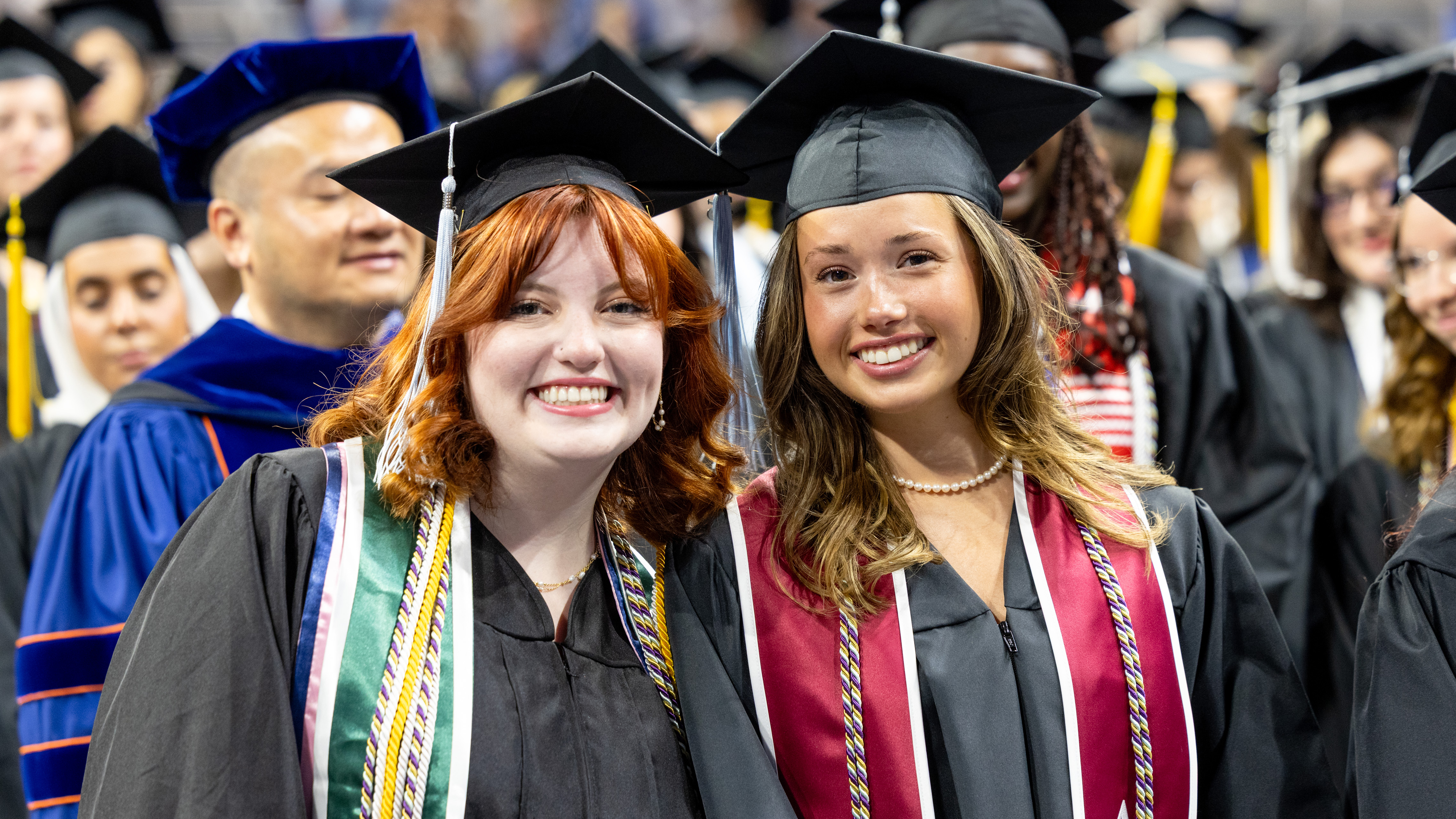 Two Drake students in caps, gowns, and honors sashes smile and stand close during commencement