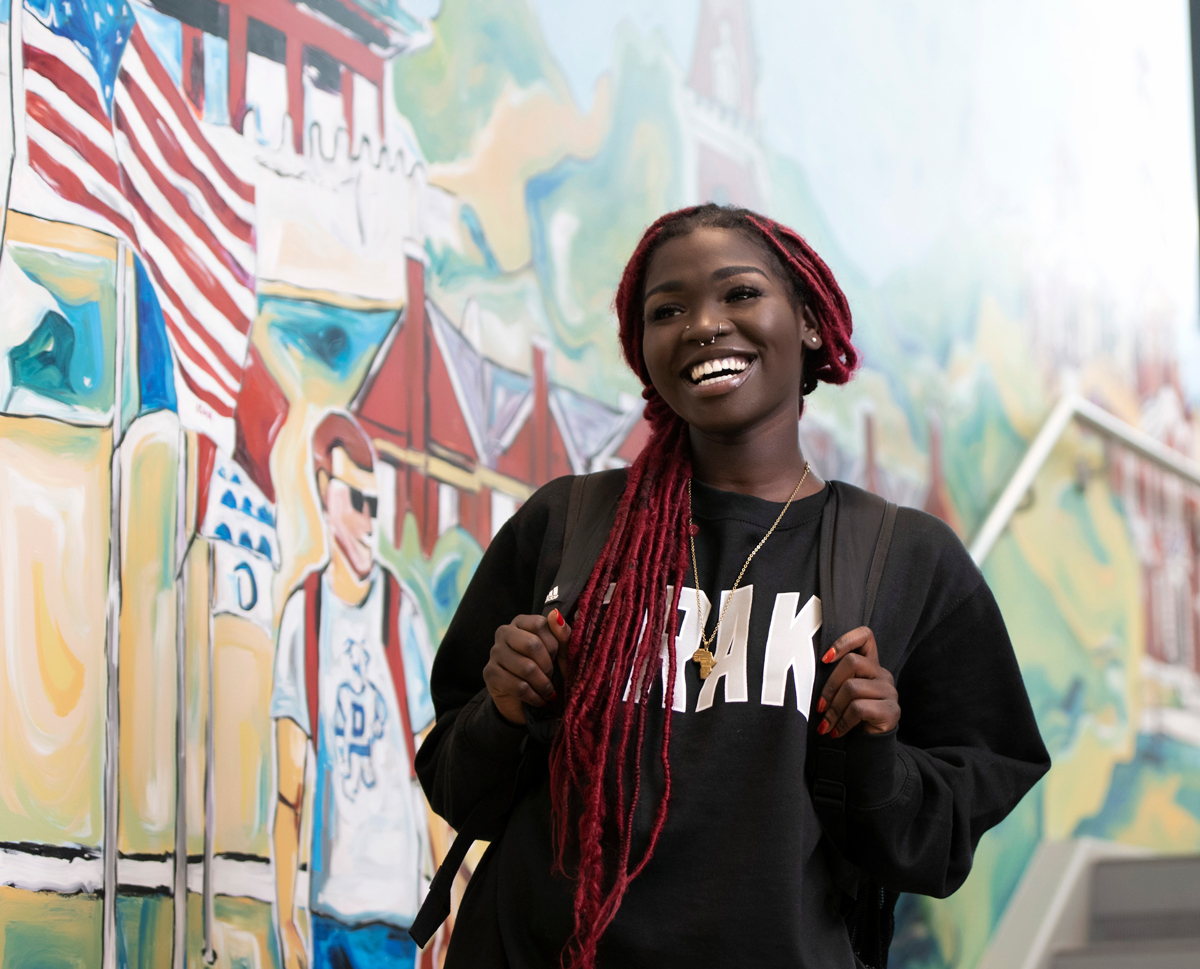 A student stands smiling in front of a wall mural