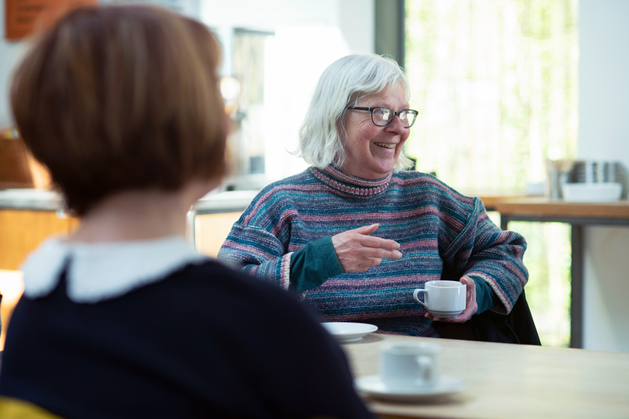An elderly woman sits, smiling and conversing with others while holding a tea cup