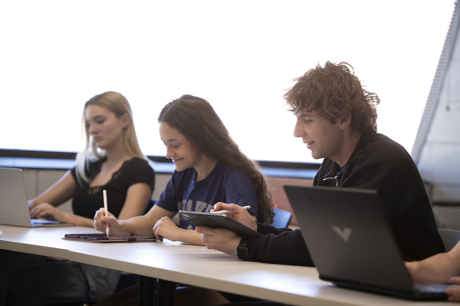 Three students sit at a table in front of laptops and notebooks.