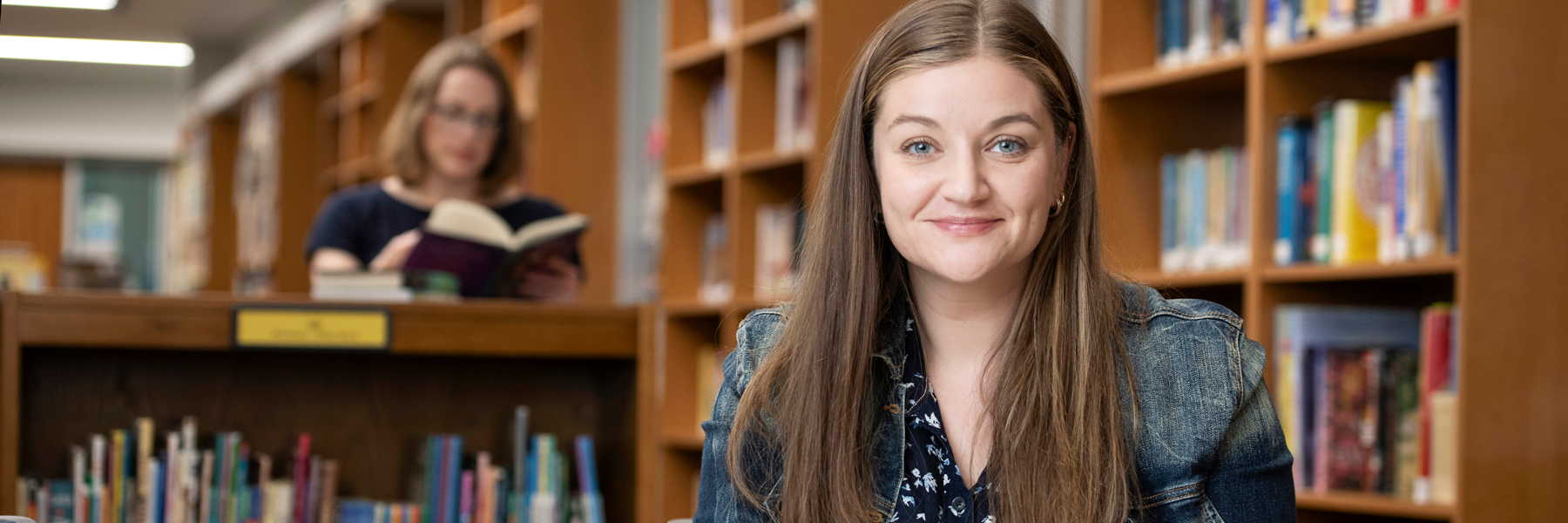 A student smiles at the camera while sitting at a table in a library with shelves of books in the background