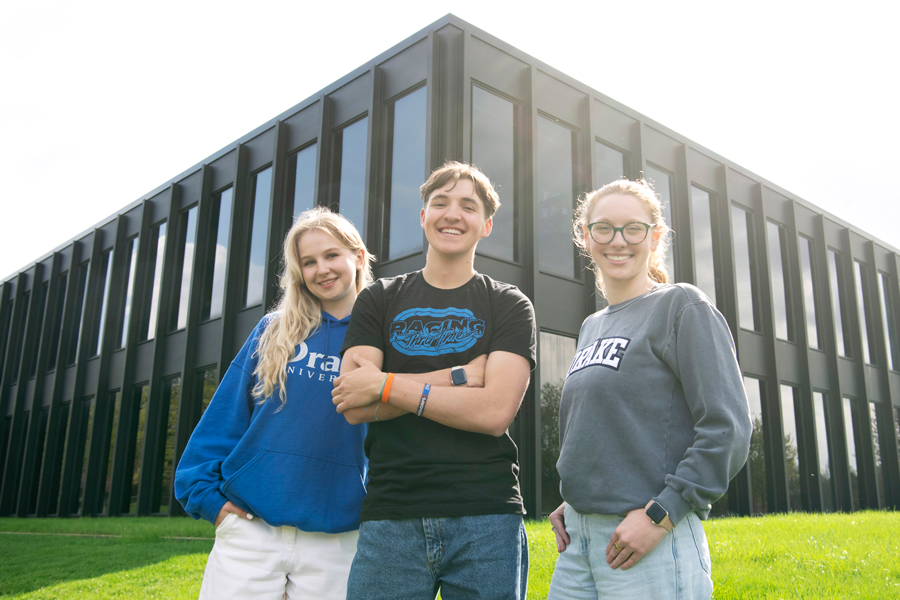 Three student stand smiling in front of a building exterior on campus