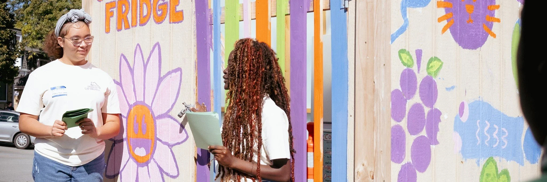 Two student stand while holding papers in front of a brightly colored outdoor mural