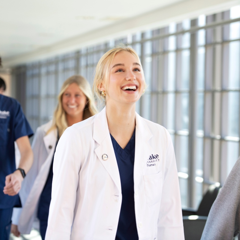 A Drake student in a white lab coat smiles and looks ahead while walking through the College of Pharmacy and Health Sciences