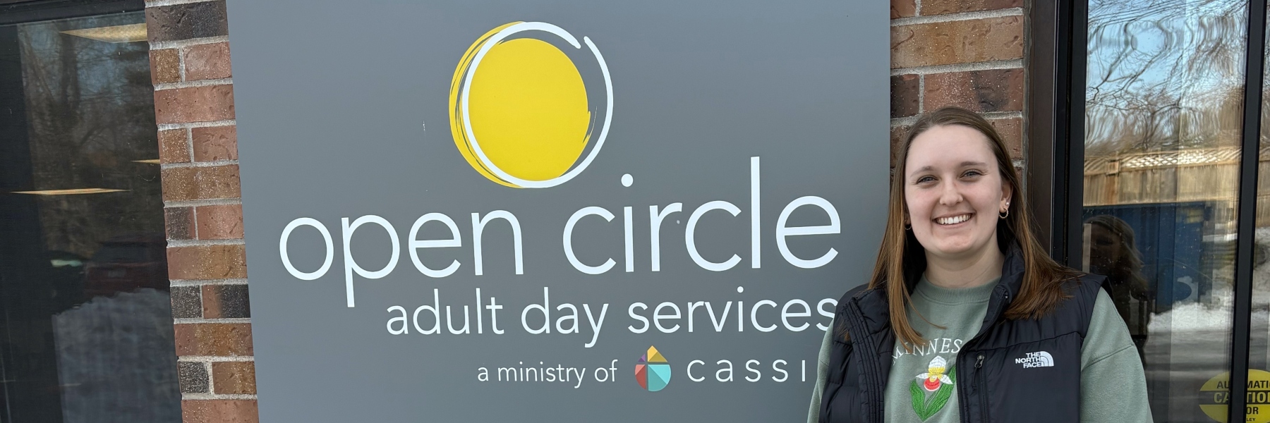 A Drake OT student smiles in front of a building with a sign saying "Open Circle Adult Day Services"