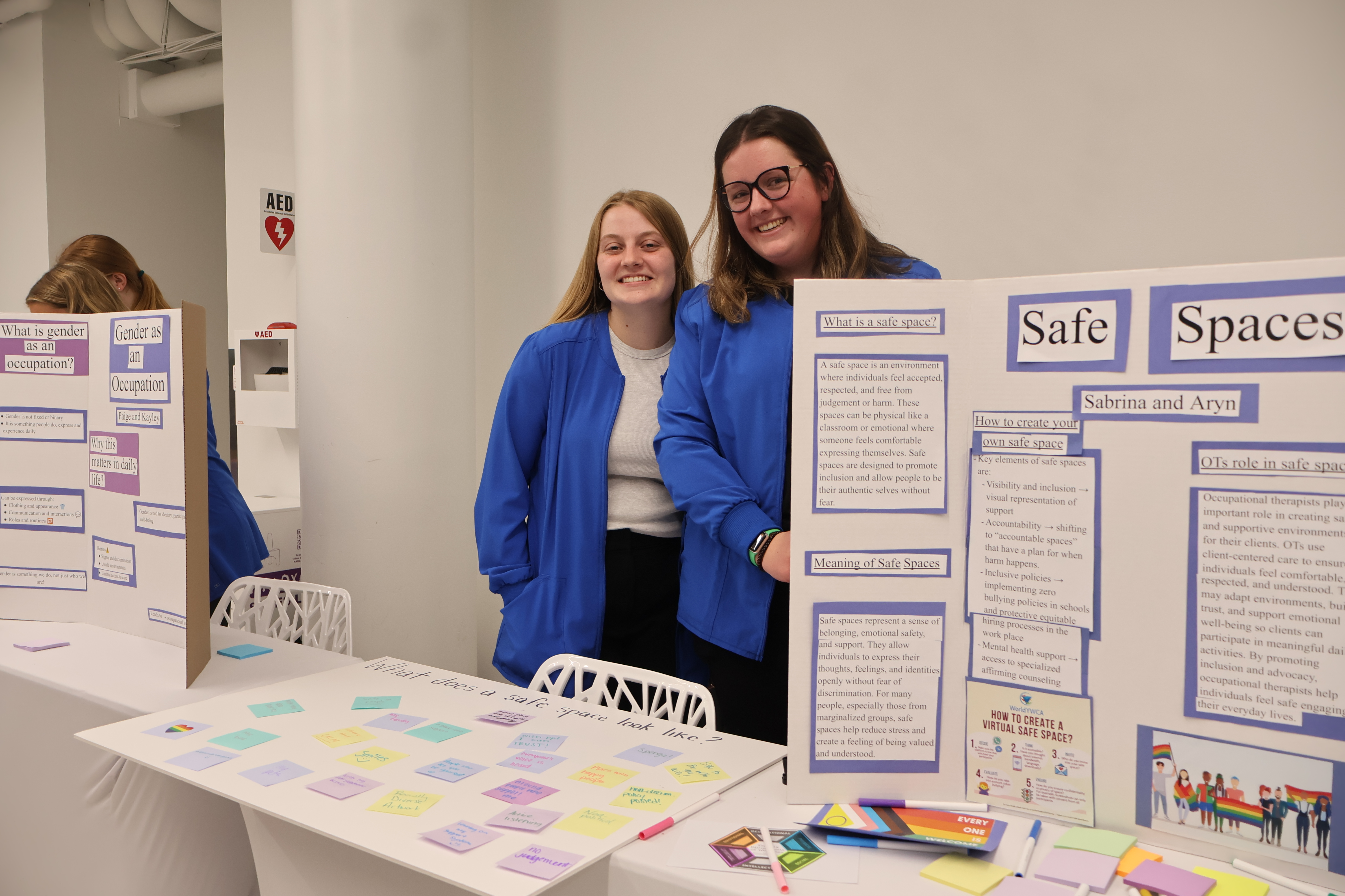 Drake OT students smile in front of their booth with a Safe Spaces poster and rainbow pride flags
