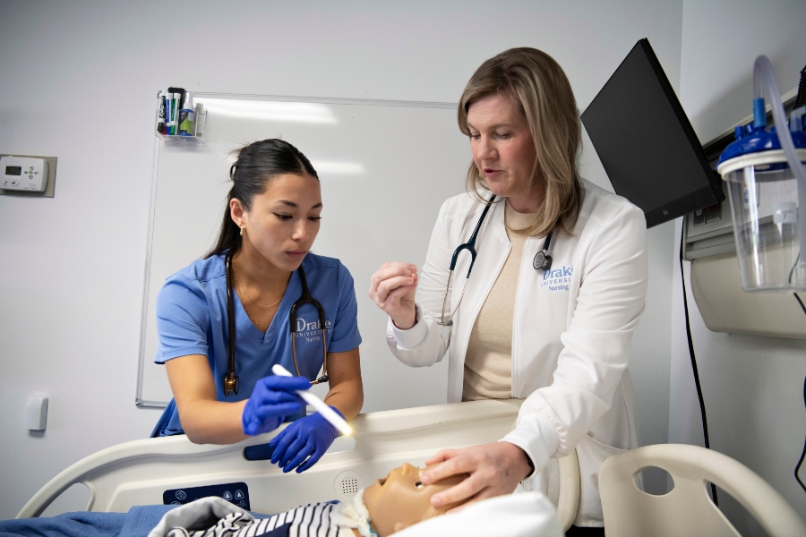 A Drake student wearing scrubs shines a light into the eyes of a mannequin while being instructed by a professor