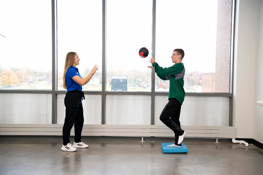 A student standing on one foot on a platform throws a ball to another student