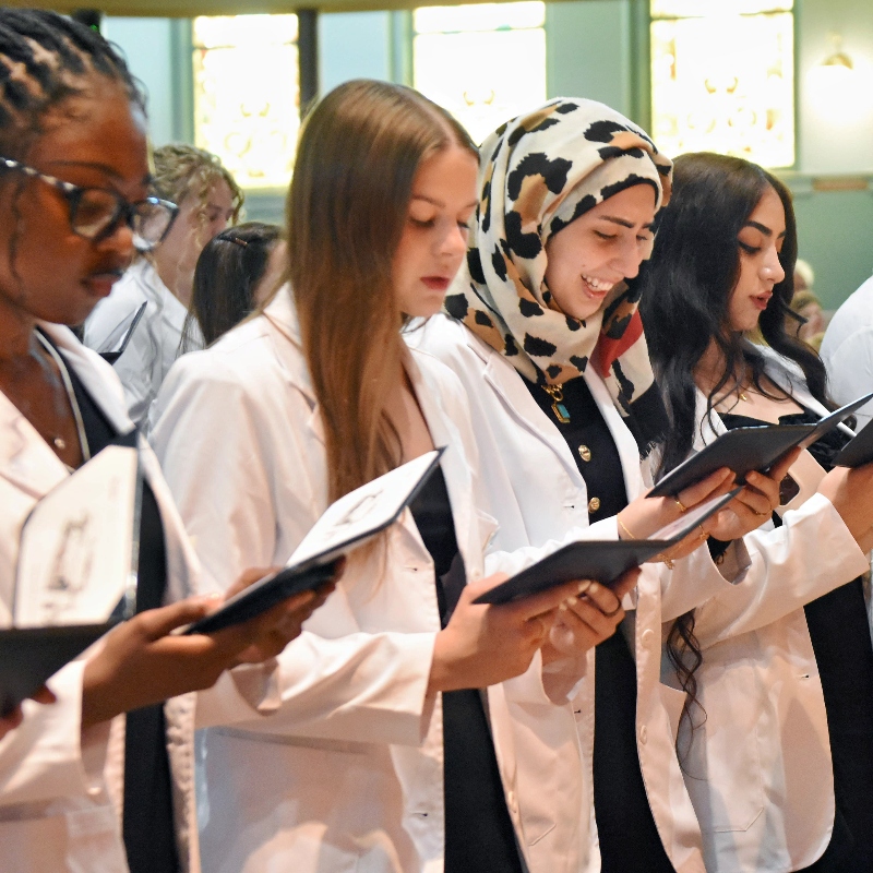 Students wearing white coats stand to recite the pledge of professionalism at their white coat ceremony