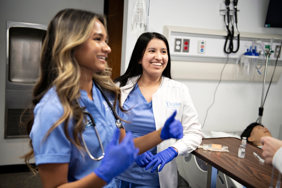 Two Drake students in scrubs and white coats smile in an exam room