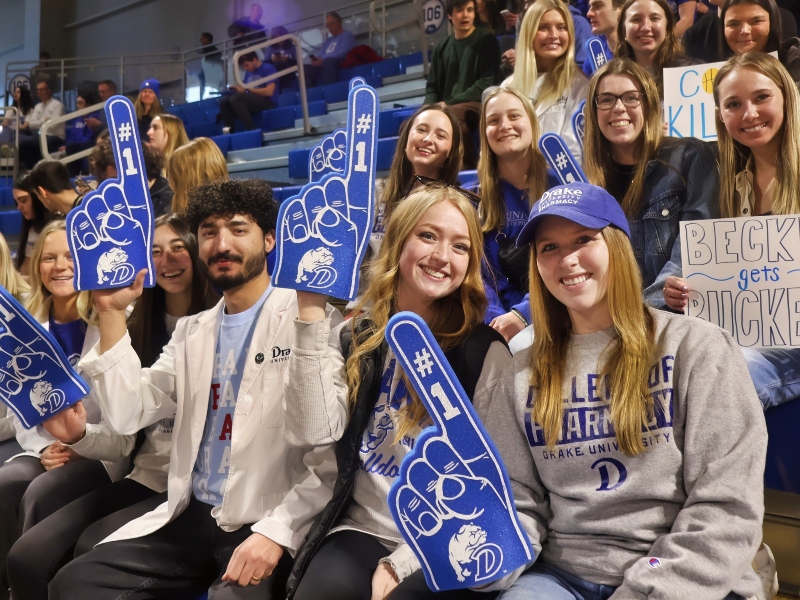 Drake CPHS students hold up foam fingers and cheer in the bleachers at a Drake basketball game