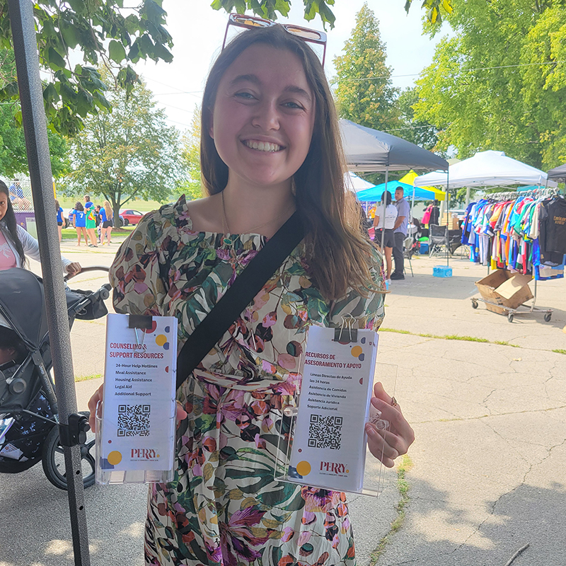 Smiling woman at an outdoor community event holding two bilingual counseling and support resources cards with QR codes.
