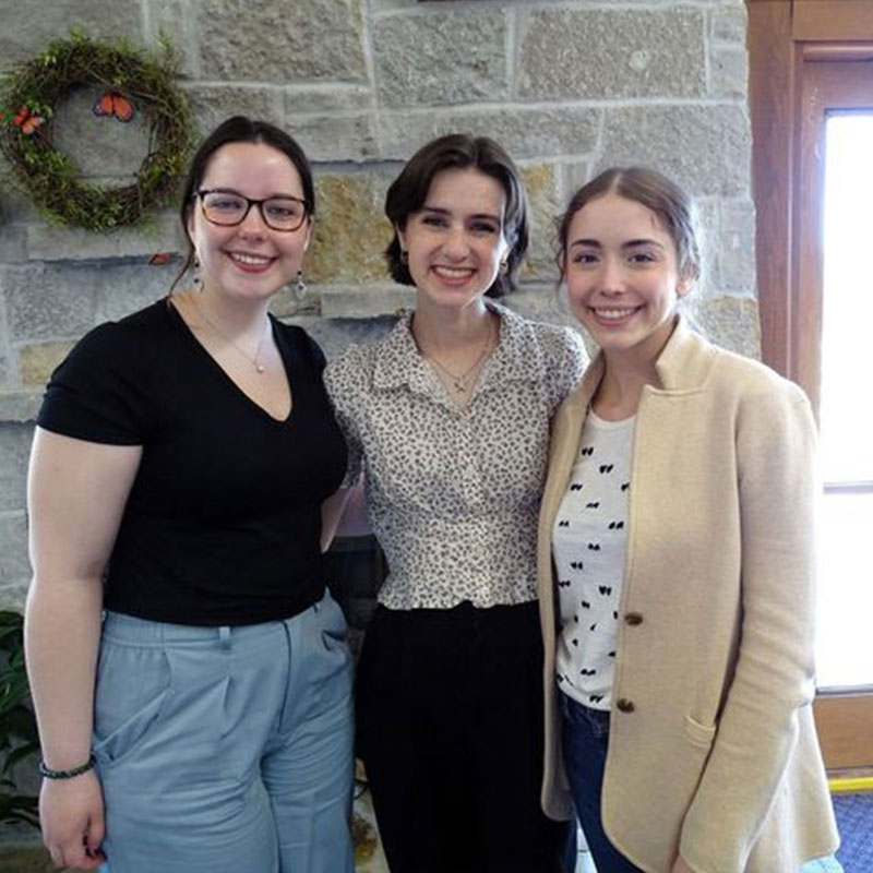 Three smiling women standing together indoors in front of a stone wall, with a wreath decoration and a window nearby.