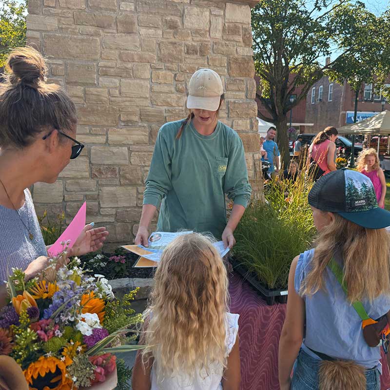 A vendor in a cap hands out papers to two children at an outdoor market stall, with flowers and potted plants in the foreground.