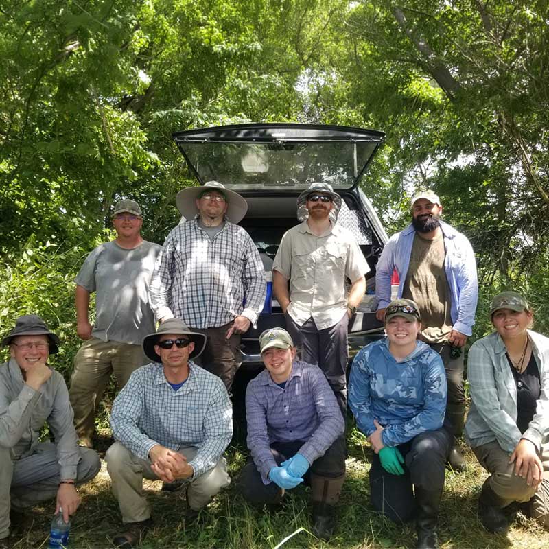 Group of nine people in outdoor work clothes pose in a wooded area in front of an open vehicle hatch.