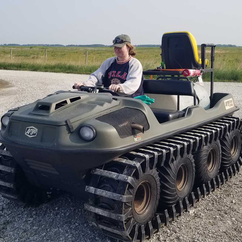 Person driving an all-terrain utility vehicle with tank-like tracks on a gravel road beside open fields.