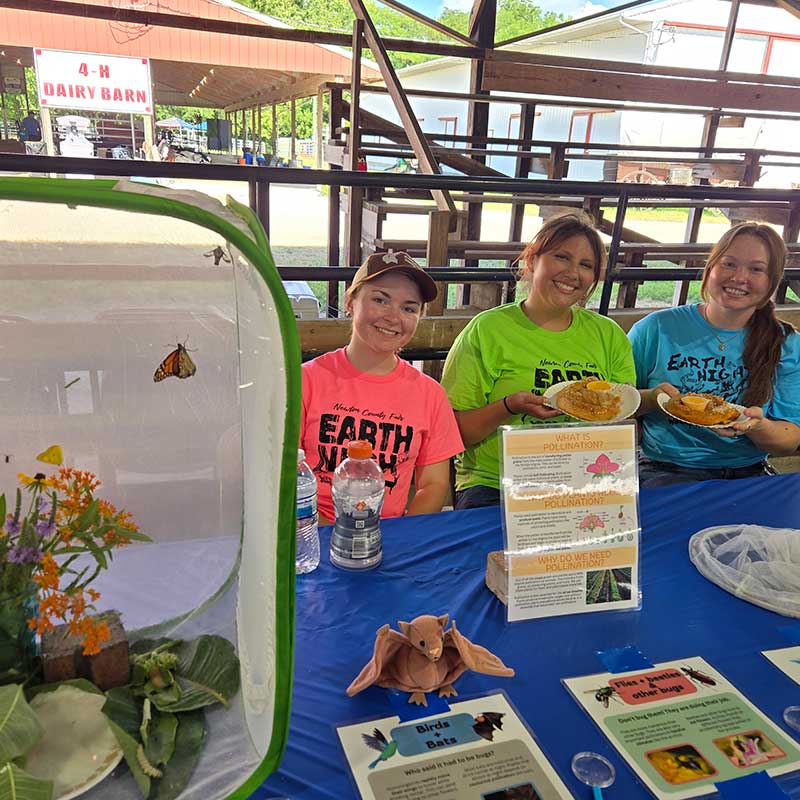 Three people at a booth sit behind a table with pollination information, craft bats, and a butterfly habitat display, holding plates of food in a barn setting.