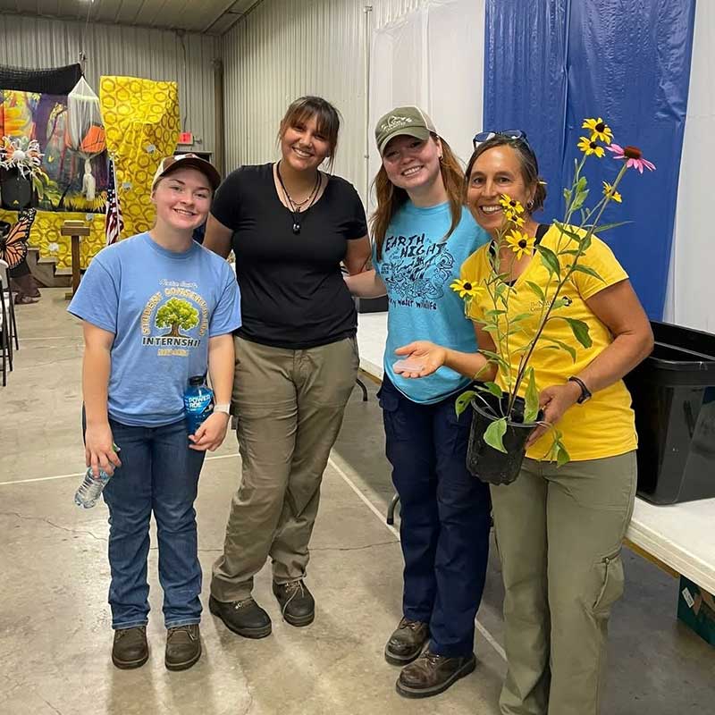 Four people smiling indoors at an event, with one holding a potted wildflower plant with yellow and pink blooms.