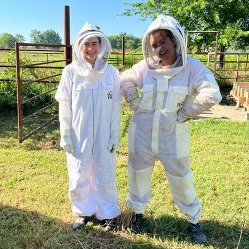 Two people wearing white beekeeping suits with veiled hoods stand smiling in a sunny grassy field near a metal fence.